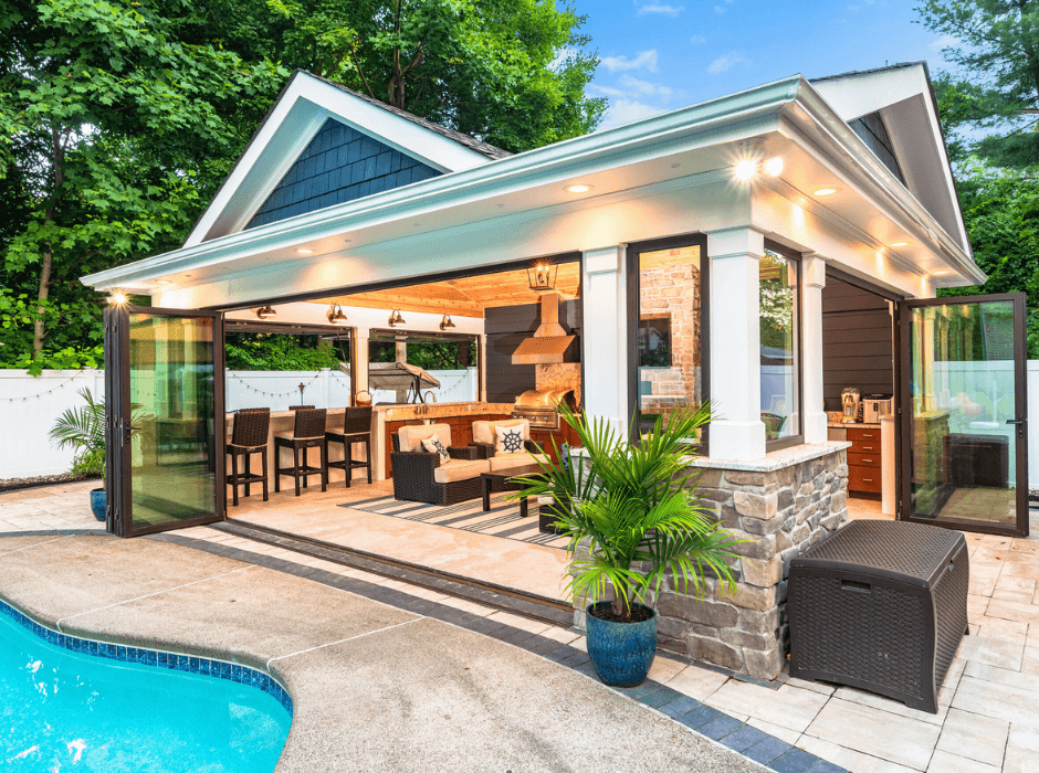 ACTIVWALL Outdoor living space with a pool, featuring a covered area with an open kitchen, bar seating, a couch, plants, and sliding glass doors. The area is surrounded by greenery and a white fence.