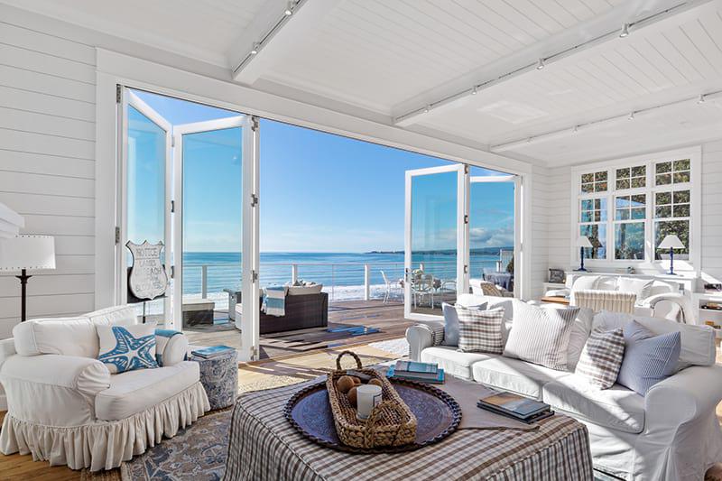 AG Millworks Spacious beach house living room with white furniture, light blue accents, and large open glass doors leading to a wooden deck overlooking the ocean.