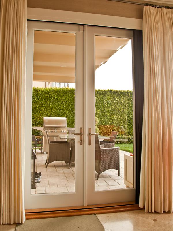 AG Millworks(swing) A set of white-framed glass patio doors with beige curtains opens to an outdoor patio area with wicker furniture and a green hedge.