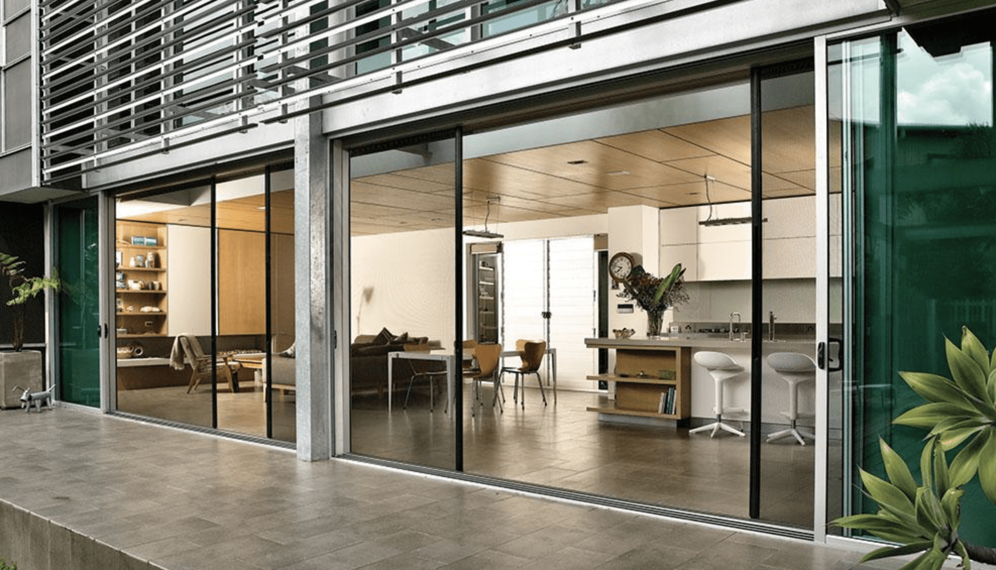 Centor(screens) Modern open-plan living area with floor-to-ceiling sliding glass doors, showcasing a living room, kitchen, and dining space with contemporary furnishings and wooden accents.