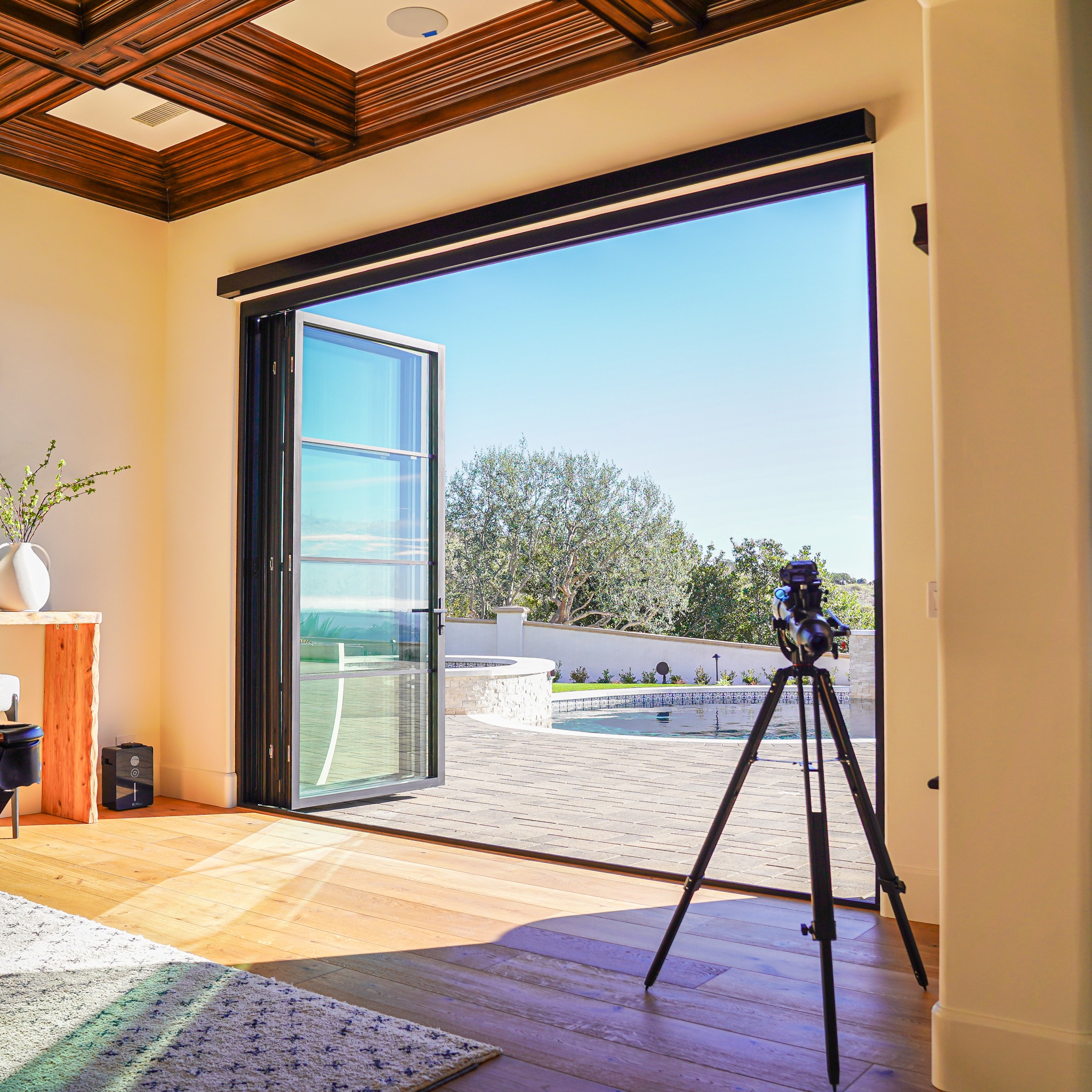 Lacantina A camera on a tripod is set up at an open patio door, overlooking a terrace with a pool and trees beyond, shot from inside a modern room with wooden floors and ceiling.