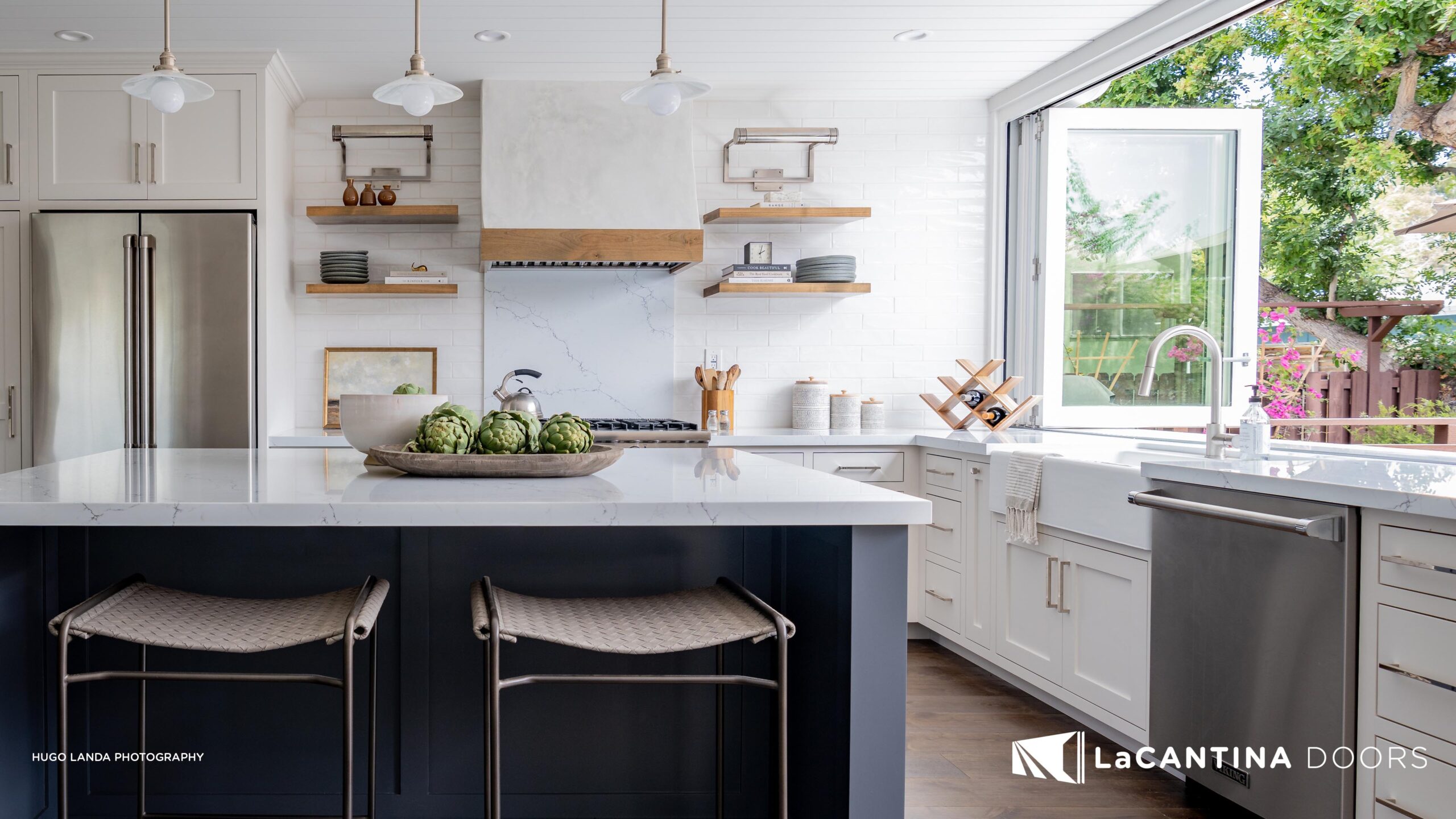 3840x2160_LCD_imagery_lowres_39 Modern kitchen with white cabinetry, an island with a dark blue base and gray bar stools, stainless steel appliances, and a large open window showcasing a garden view. LaCantina Doors branding visible.