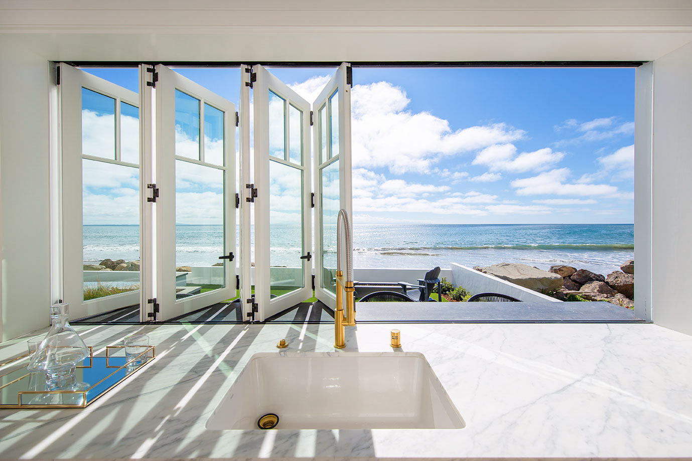 AGM-Bifold-window-09 A kitchen sink with a marble countertop is positioned in front of open windows offering a clear view of a sunny beach and ocean.