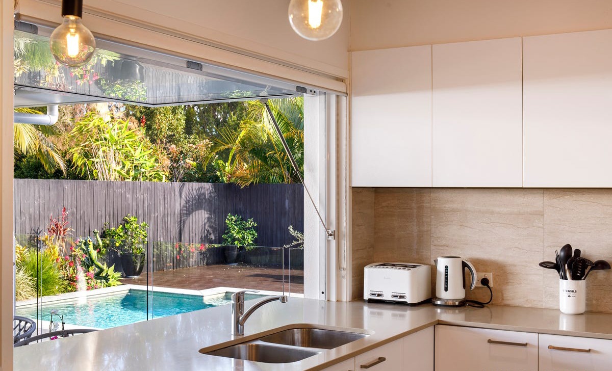 gravity window A modern kitchen with white cabinets and a sink overlooks a backyard with a swimming pool. An electric kettle and a toaster are on the counter. The backyard features lush greenery and a wooden fence.