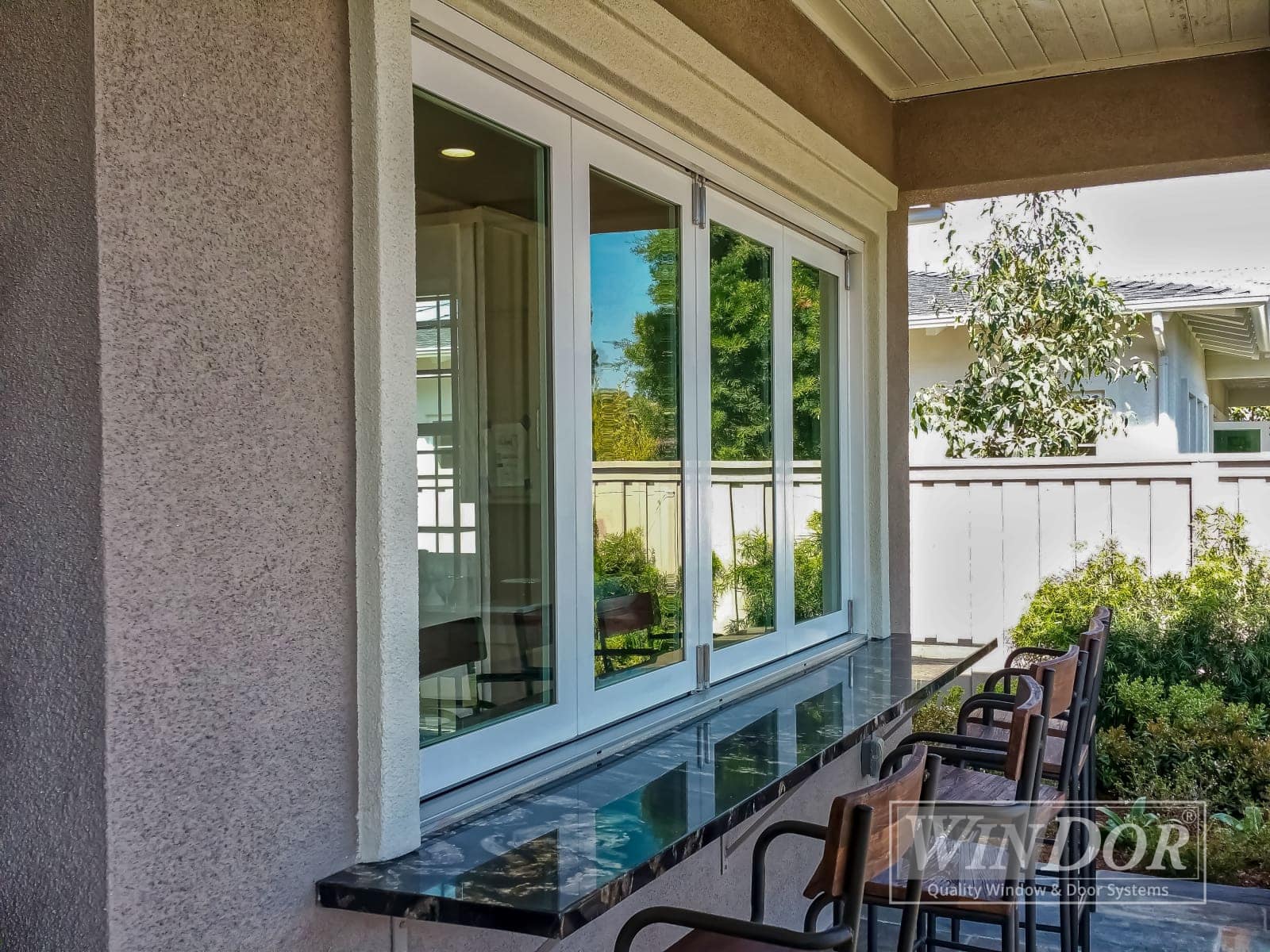 windorwindow1 A large sliding window installed on the exterior wall of a house, with three barstools placed in front. The window frame is white, and there is a logo in the bottom right corner that reads "WIN-DOR.