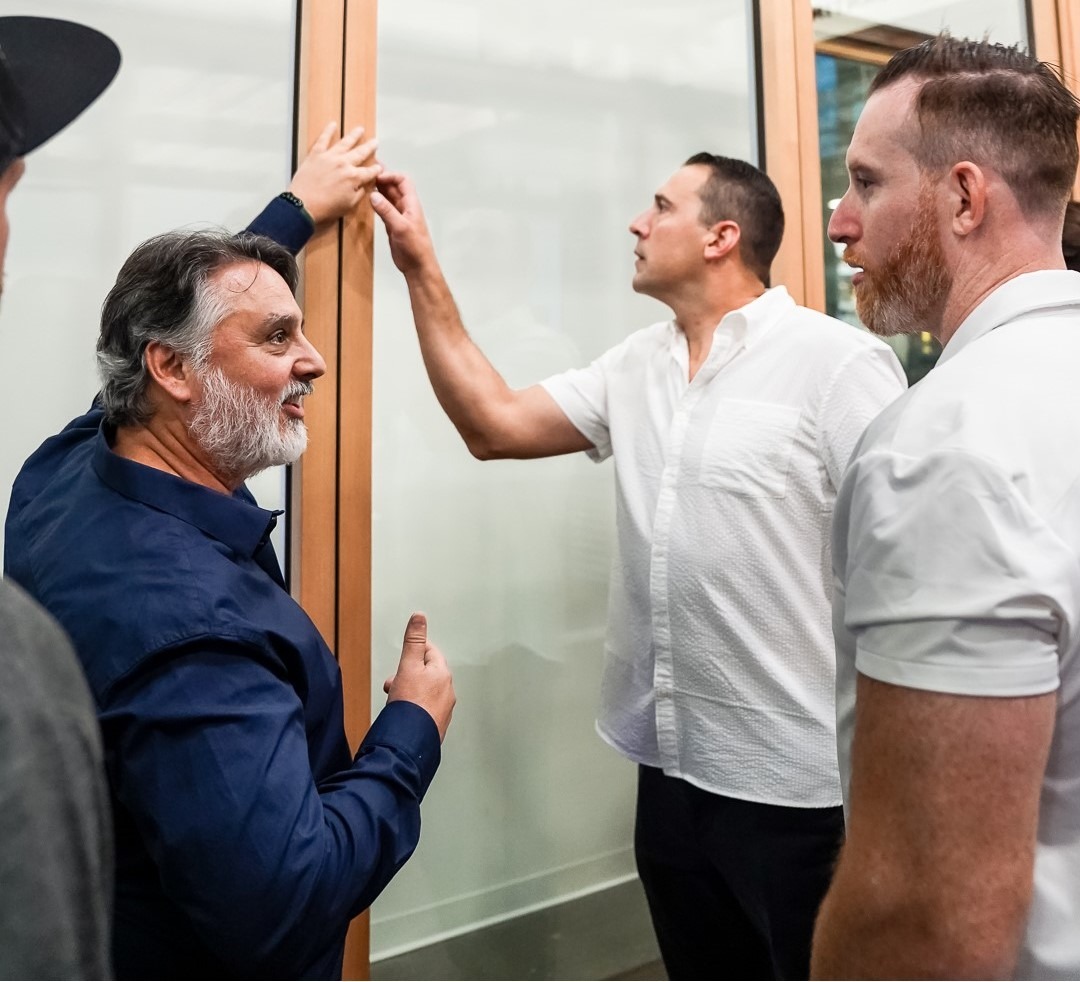 LUXEEvent4 Three men engage in a discussion around a glass door, with one man pointing and another examining the frame closely.
